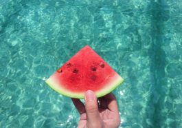 hand holding a slice of watermelon with blue swimming pool water in the background