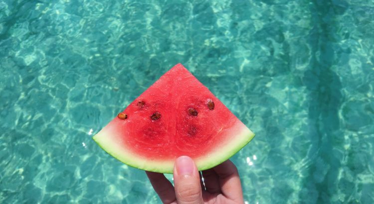 hand holding a slice of watermelon with blue swimming pool water in the background
